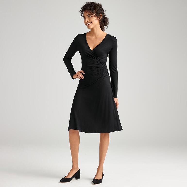 Frau in schwarzem Kleid mit langen Ärmeln, modisch in einem hellen Raum stehend.