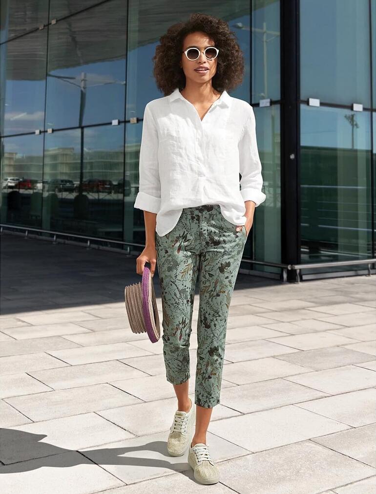 Frau in einer weißen Leinen-Tunika und gemusterten Hosen steht lächelnd im Freien.