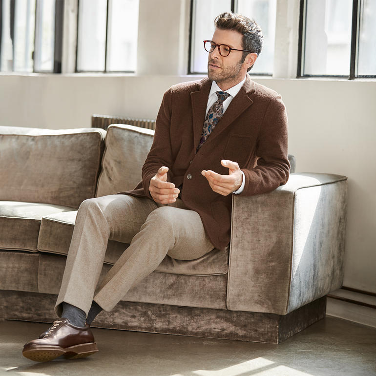 Mann trägt braunen Blazer mit einem gestreiften Krawattenmuster, sitzt auf einem Sofa.