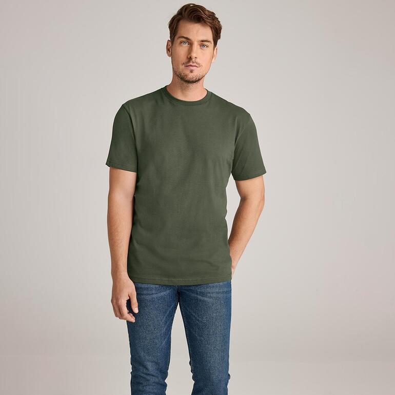Mann in olivgrünem T-Shirt mit Jeans, stehend vor neutralem Hintergrund.