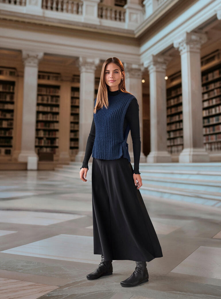 Frau in einem dunkelblauen Alpaka-Pullunder, vor Regalen in einer Bibliothek.