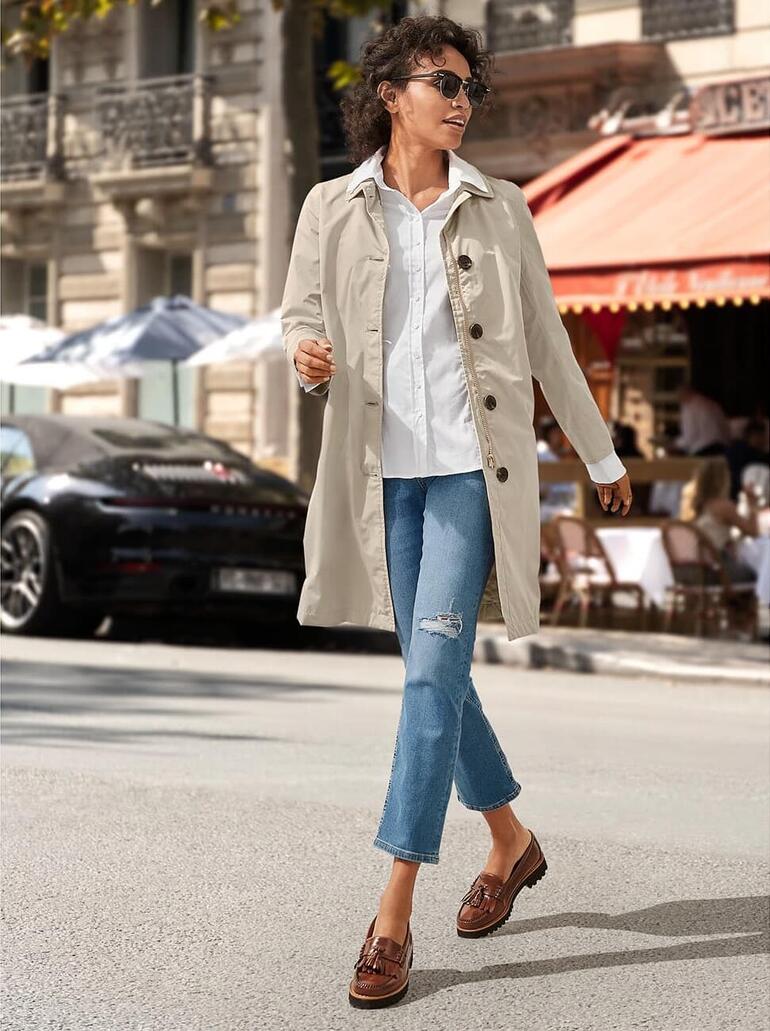 Frau in beigem Trenchcoat und weißen Sneakers, die durch eine belebte Straße mit Cafés geht.