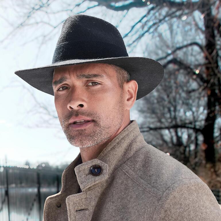 Mann mit schwarzem Alpaka-Hut und grauem Mantel, im Freien.