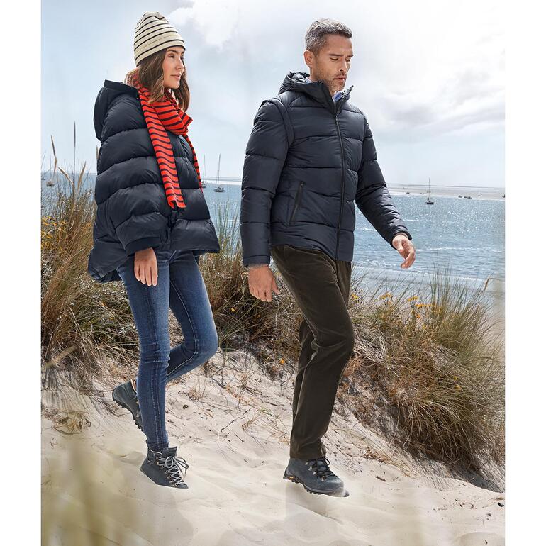 Paar in Winterkleidung, darunter eine Recycling-Daunenjacke, spaziert am Strand.