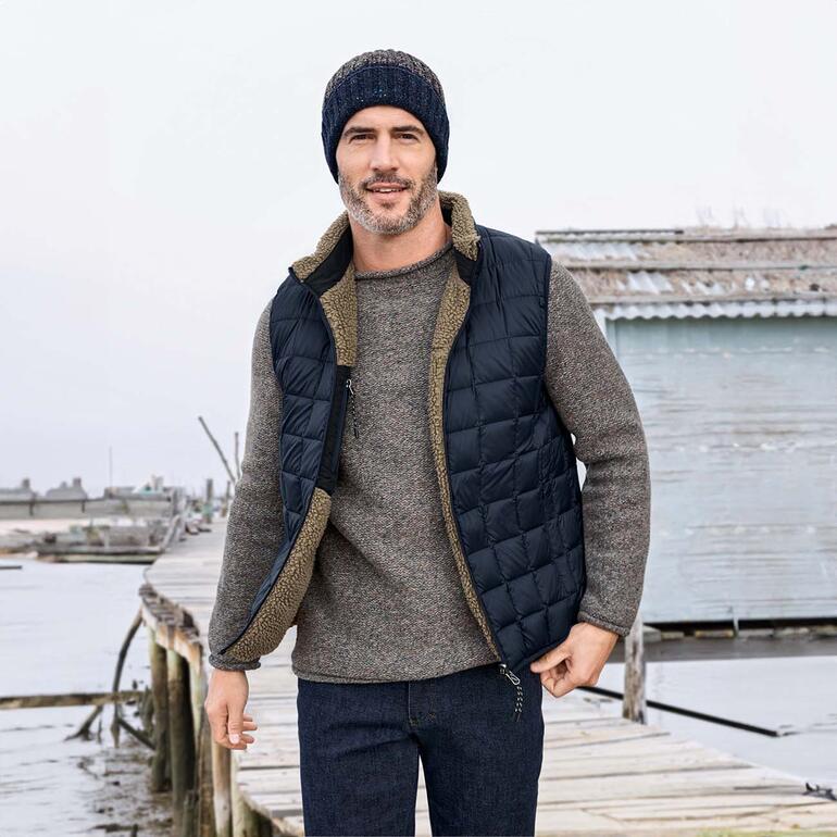 Mann in Daunen-Wendeweste posiert am Hafen mit grauem Pullover und Mütze.
