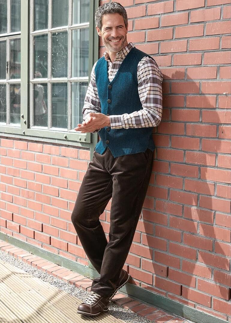 Mann in blauer Weste mit kariertem Hemd an Ziegelwand.