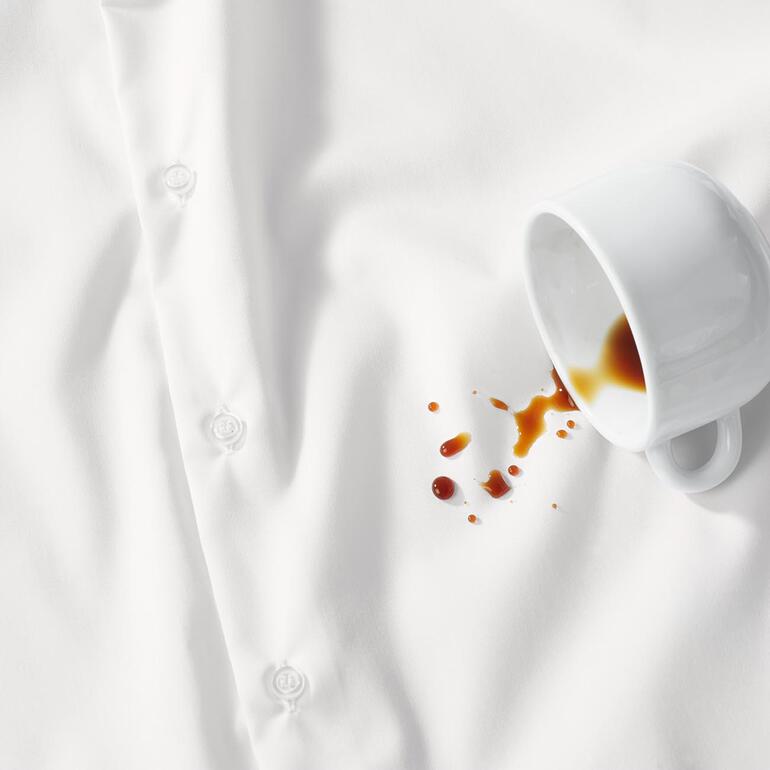 Dank Nano-Technologie perlen Flüssigkeiten wie Kaffee oder Rotwein ab, und Schmutz lässt sich mit Wasser mühelos entfernen. Weißes Hemd mit Kaffeefleck und Tasse daneben.