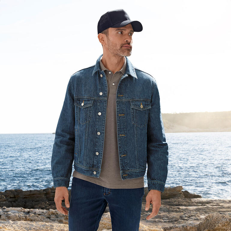 Stetson Trucker-Cap Mann in OCONI Hanf-Jeansjacke steht am Meer mit einem grauen Polohemd.