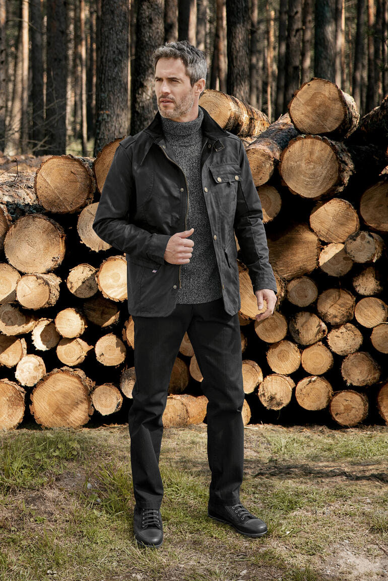 Mann in Wachsbaum-Jacke steht in einem Wald vor Baumstämmen.