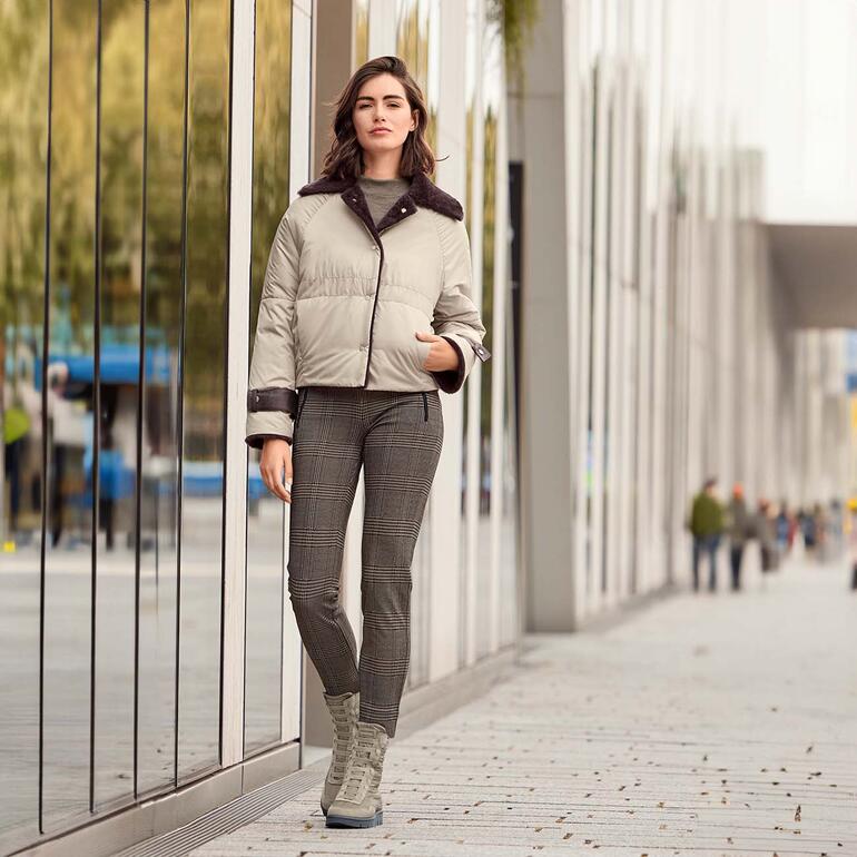 Frau in beiger Lammfell-Wendejacke steht vor modernen Gebäuden.