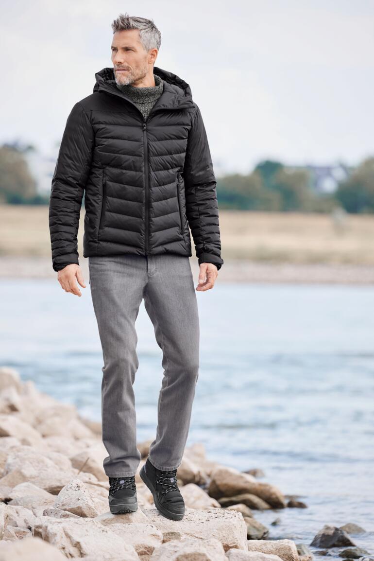 Mann in Daunen-Regenjacke, steht am Wasser und lächelt.