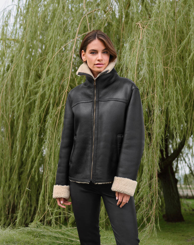 Frau trägt schwarze Curlylamm-Fliegerjacke vor einem grünen Weidenbaum.