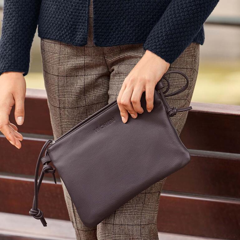 Frau in beige Jacke hält eine braune Crossbody-Tasche.