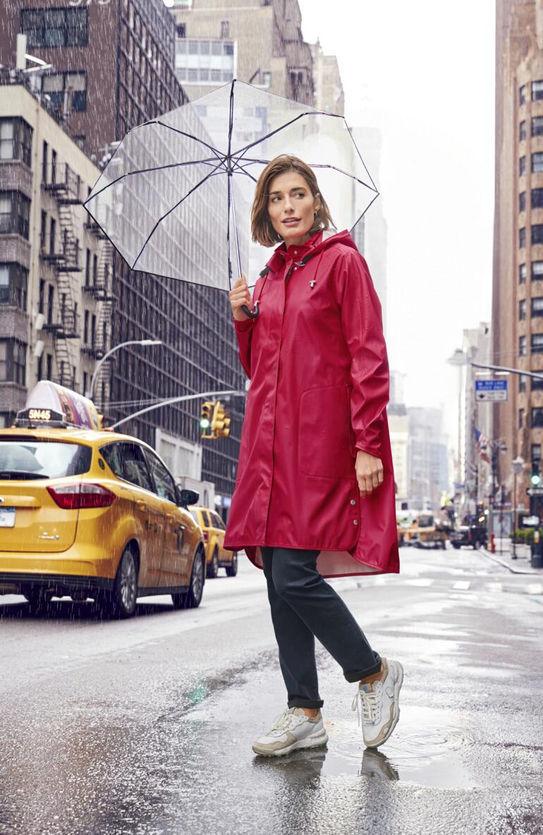 Frau in rotem Regenmantel hält einen durchsichtigen Regenschirm in einer urbanen Umgebung.