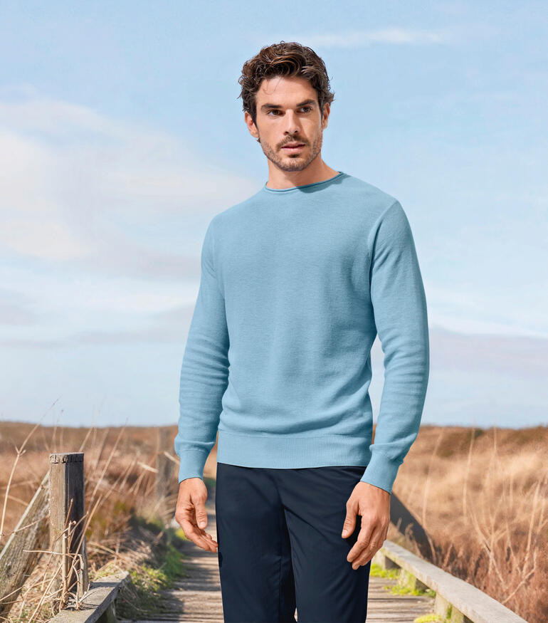Mann in einem hellblauen Edel-Piqué-Pullover steht in einer ländlichen Umgebung.
