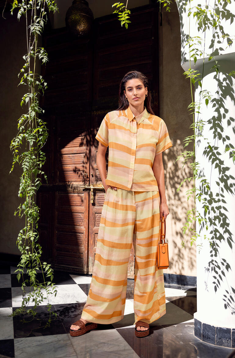 Frau im gestreiften Color-Batist-Zweiteiler mit Umhängetasche steht in einem Raum.