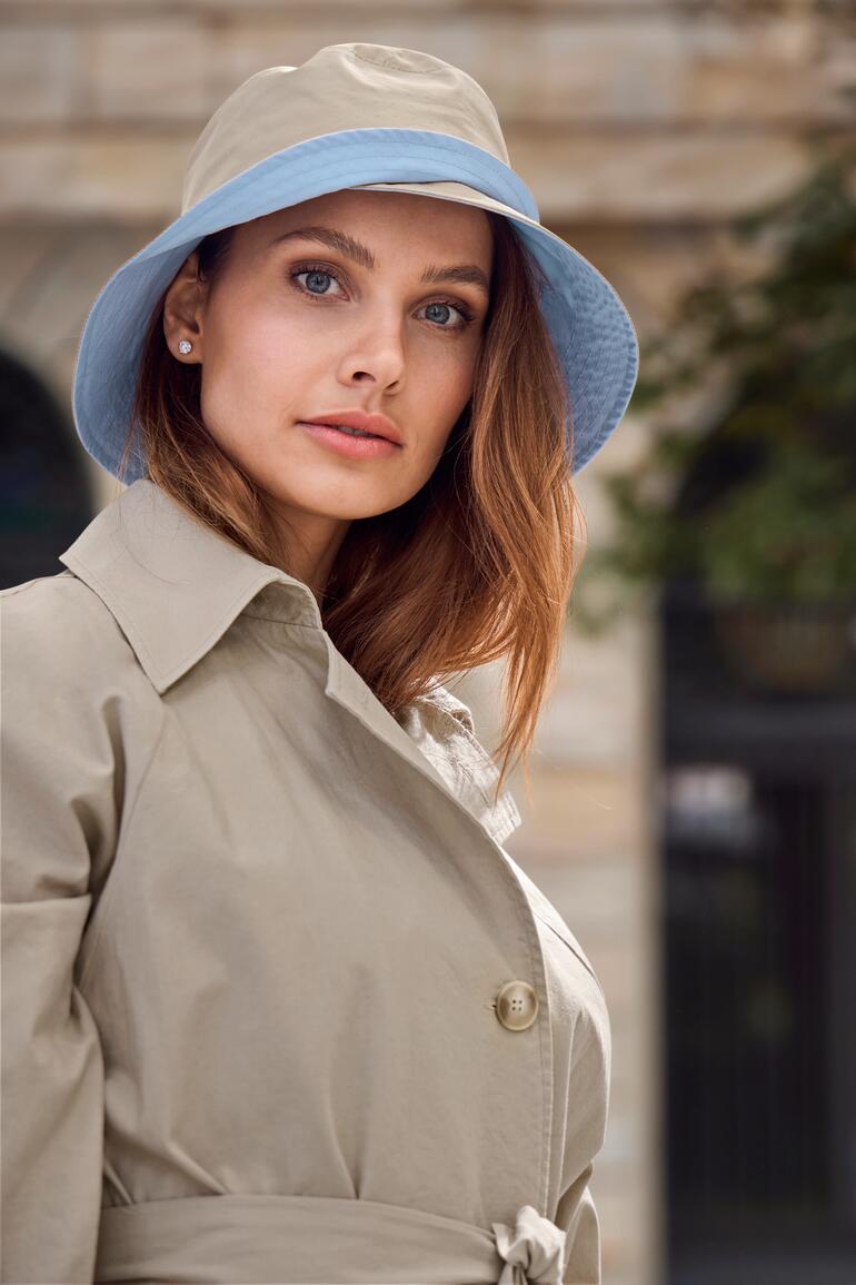 Frau trägt beigen Allwetter-Bucket-Hut und Trenchcoat, schaut in die Kamera
