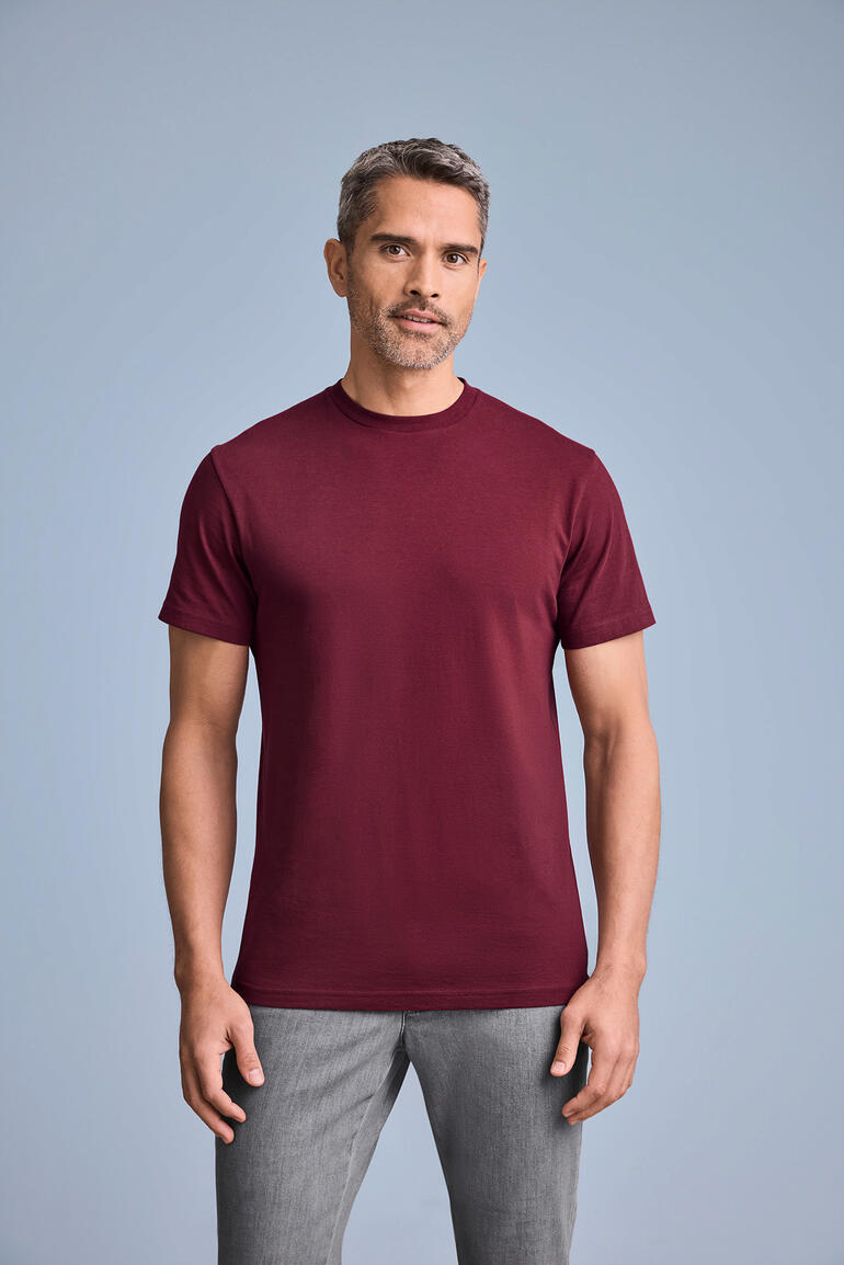 Mann trägt burdeos rotes T-Shirt mit Rundhalsausschnitt, poses vor grauem Hintergrund.