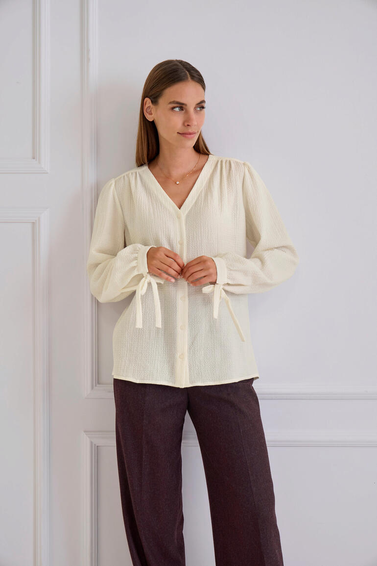 Frau steht in heller Baumwoll-Kaschmir-Bluse und braunen Hosen, an einer Wand.