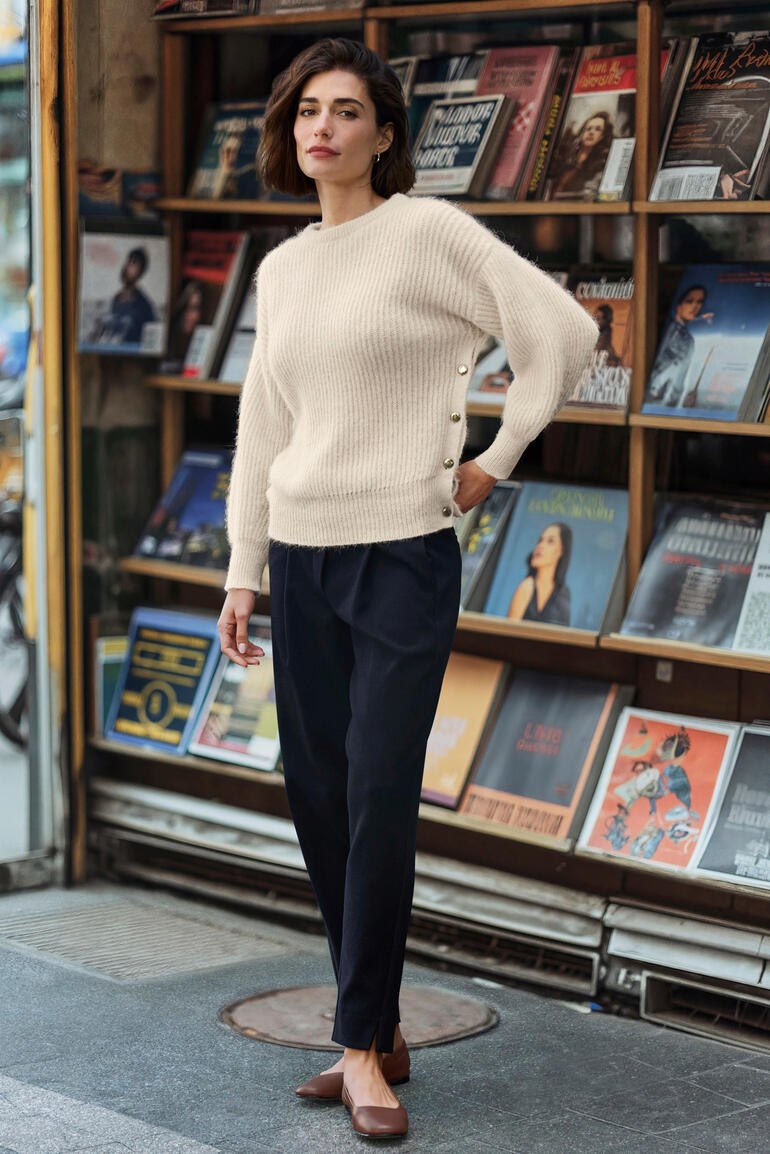 Frau steht vor einem Kiosk mit Zeitschriften, trägt einen beigen Pullover und blickt in die Kamera.