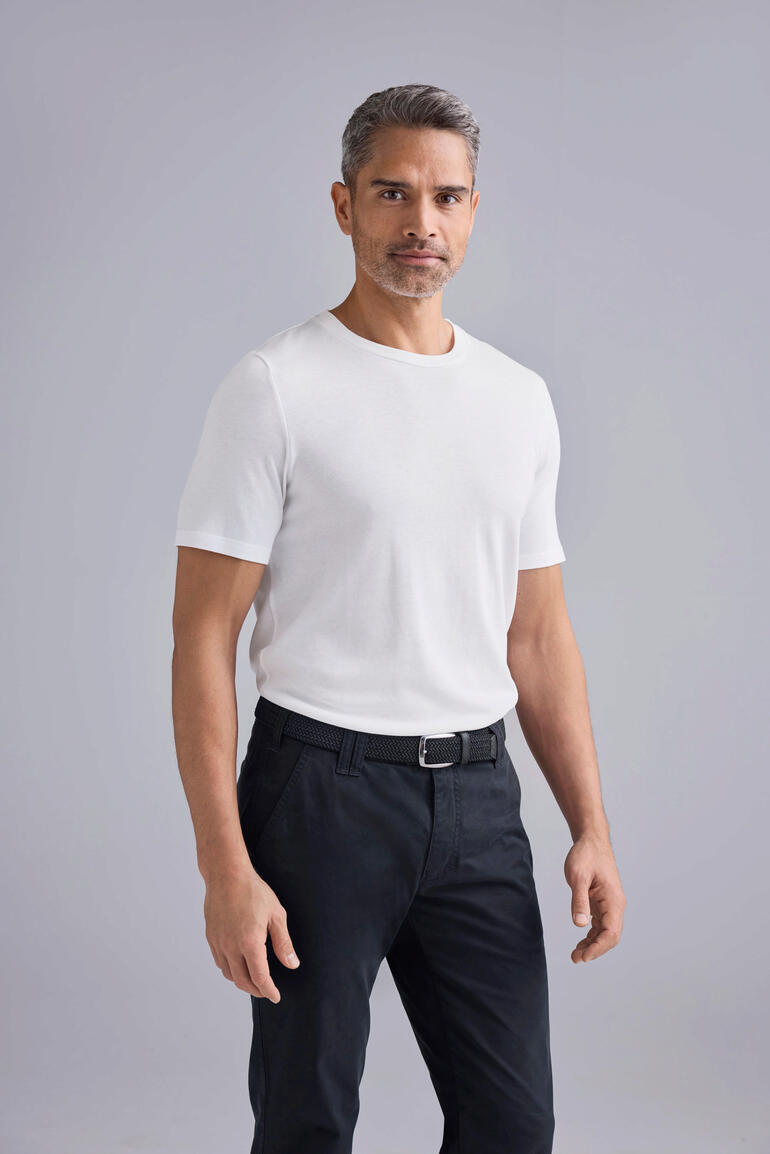 Junghans 1954 Edel-Basic-Shirt Mann in weißem T-Shirt und schwarzen Hosen, steht vor einem grauen Hintergrund.
