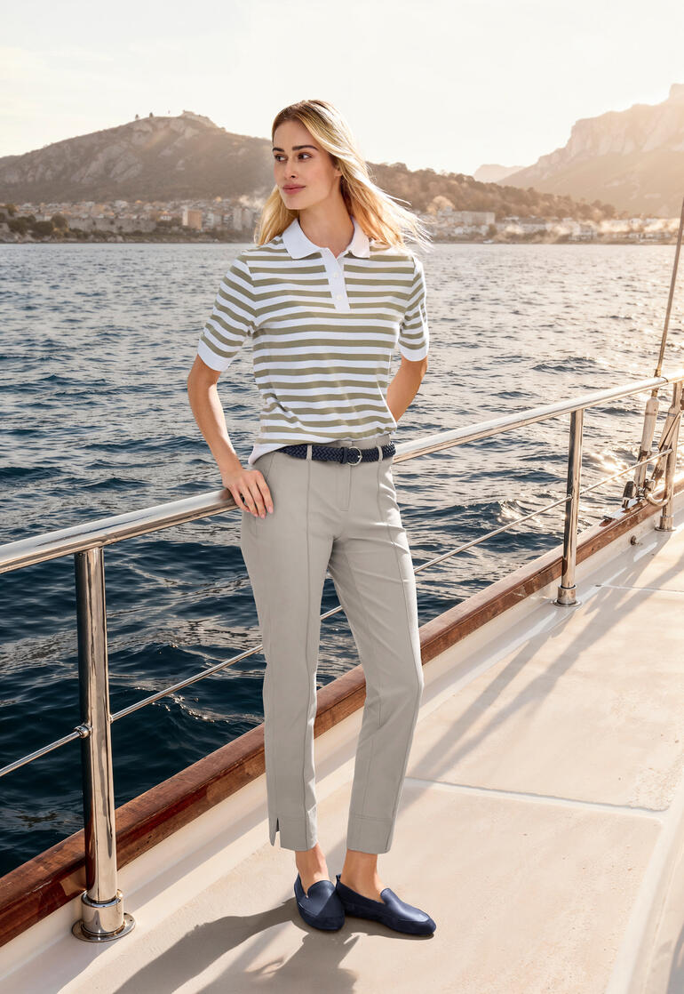 Frau trägt ein beiges Boxy-Poloshirt mit Streifen, posiert am Ufer eines Bergsees.