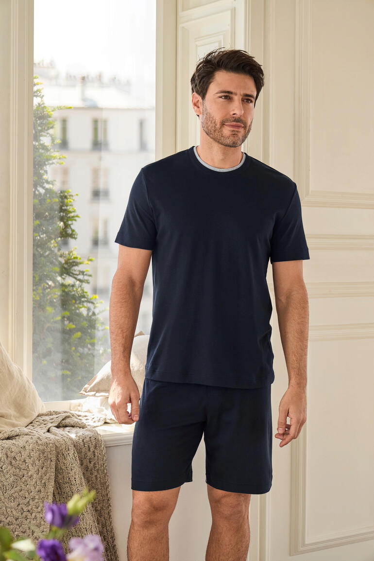 Mann steht in einem hellen Raum in einem navy-farbenen Recovery-Pyjama mit kurzen Hosen.