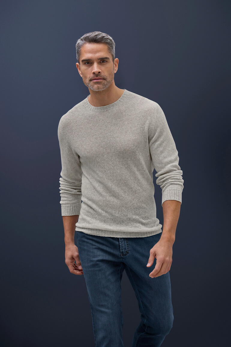 Taupe Mann trägt einen hellgrauen Kaschmir-Leinen-Pullover und Jeans vor dunklem Hintergrund.