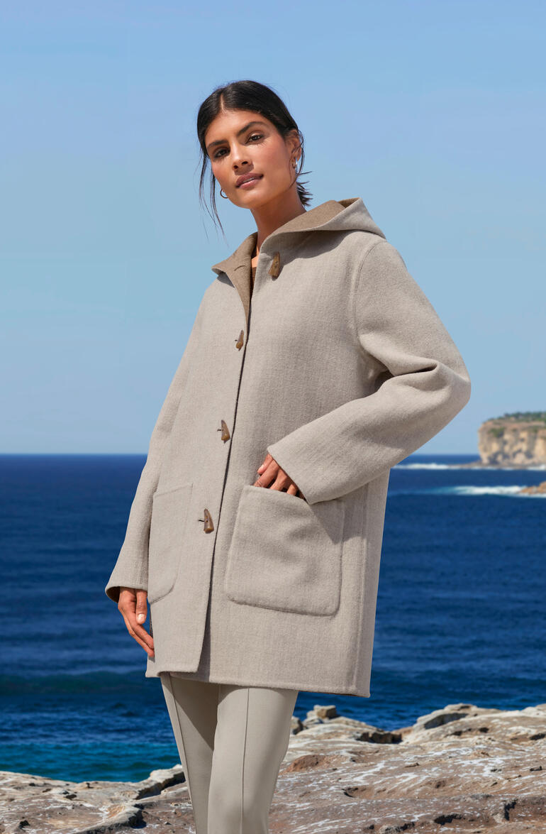 Sand/Beige Alpaka-Doubleface-Dufflecoat in Beige, getragen von einer Frau mit Meer im Hintergrund.