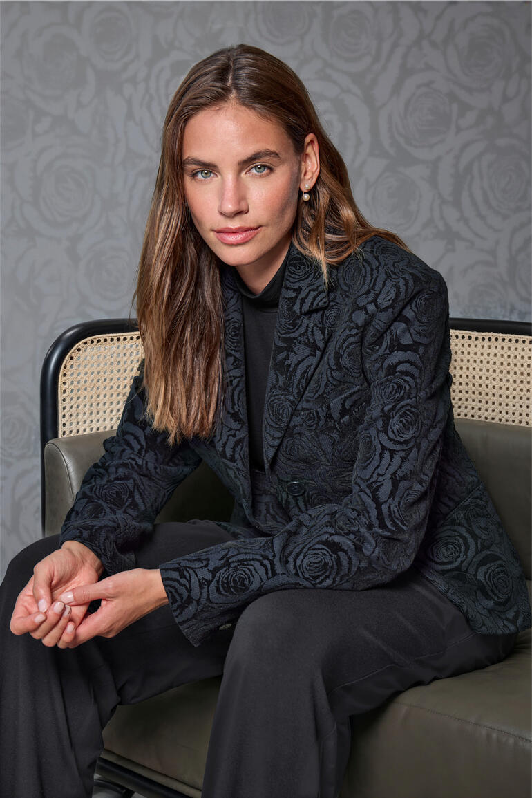 Frau in floral-gesticktem Blazer sitzt in stilvollem Raum.