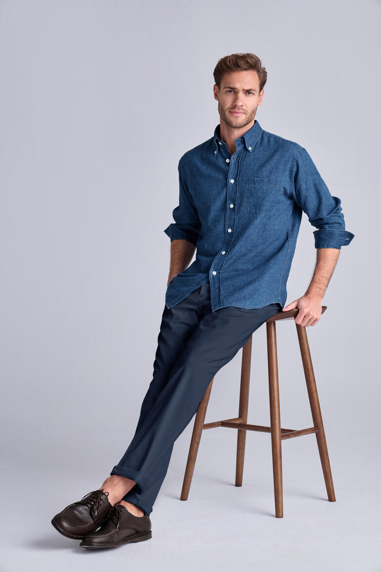 Mann sitzt auf einem Hocker und trägt ein blaues Hemd und eine dunkelblaue Chinohose.