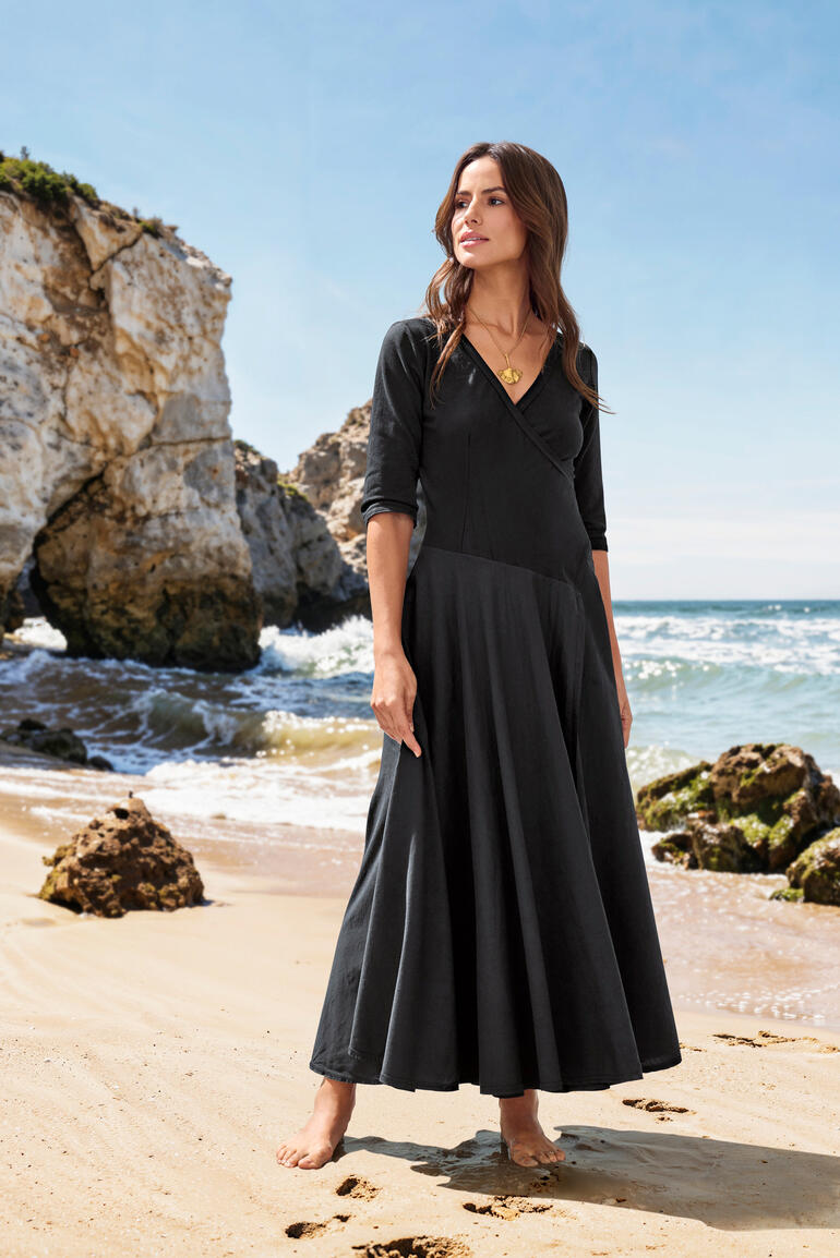 Frau im schwarzen Ibiza Kleid steht am Strand.