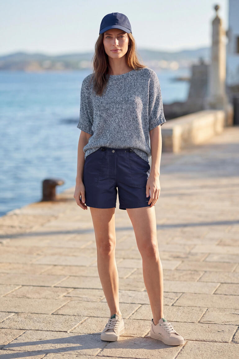 Frau steht am Meer in blauem Pulli und Shorts.