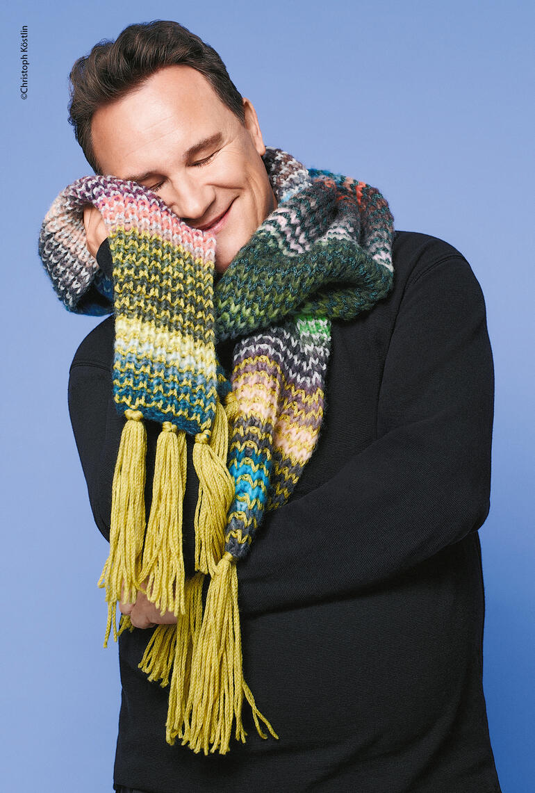 Lächelnder Mann trägt bunten Schal mit Fransen vor blauem Hintergrund.