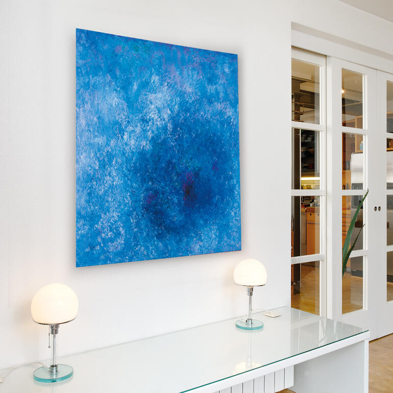 Aufgrund seiner farbigen Vielschichtigkeit entwickelt das Werk eine Eigendynamik, die jeden Raum bereichert. Abstraktes blaues Bild von Benno Werth, hängt an einer Wand über einem modernen Tisch.