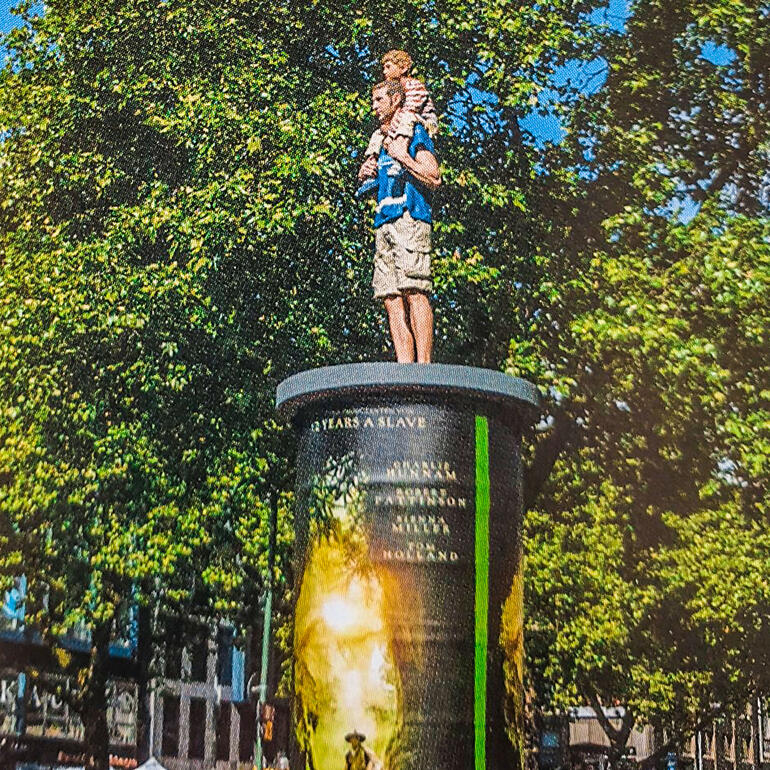 Vater und Sohn-Skulptur auf einem Platz, umgeben von Bäumen.