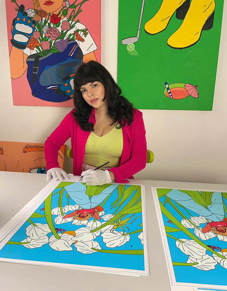 Anna Rupprecht Frau mit schwarzem Haar in pinkem Blazer sitzt an Tisch und signiert Kunstwerke.