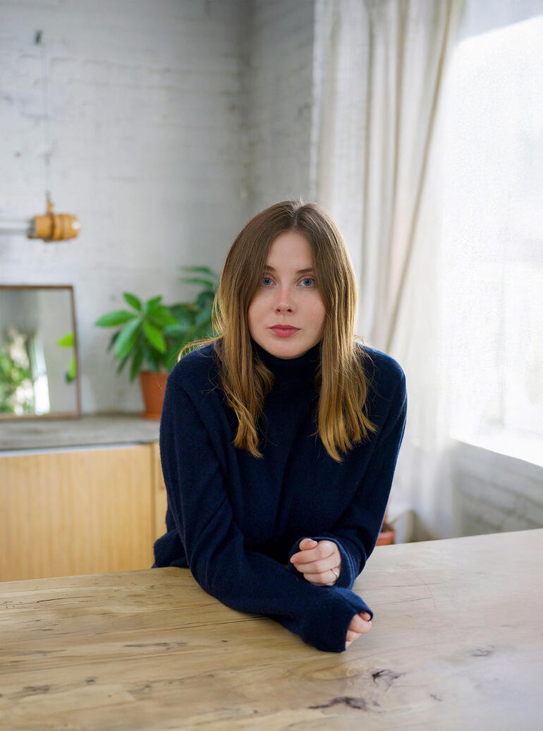 Junge Frau in einem dunkelblauen Pullover sitzt am Tisch und blickt direkt in die Kamera.