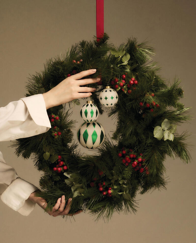 Hand mit Weihnachtskugeln über einem grünen Kranz mit roten Beeren.