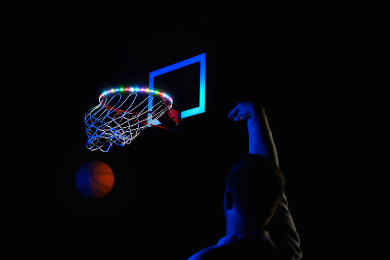 Basketballkorb mit bunten LED-Leuchten und Person beim Werfen auf einem dunklen Platz.