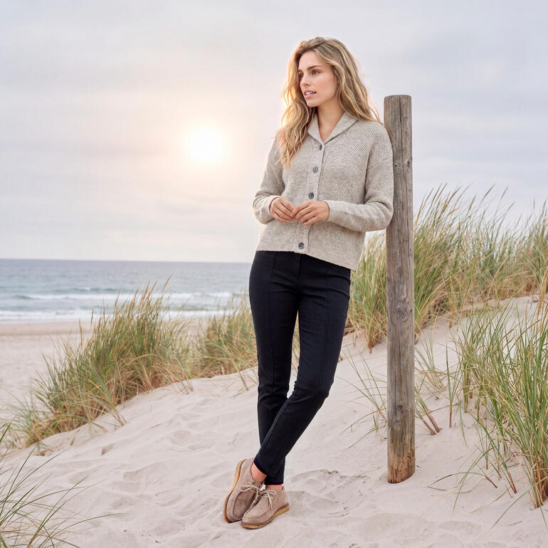 Frau in grauem Cardigan steht am Strand mit Sonnenuntergang im Hintergrund.