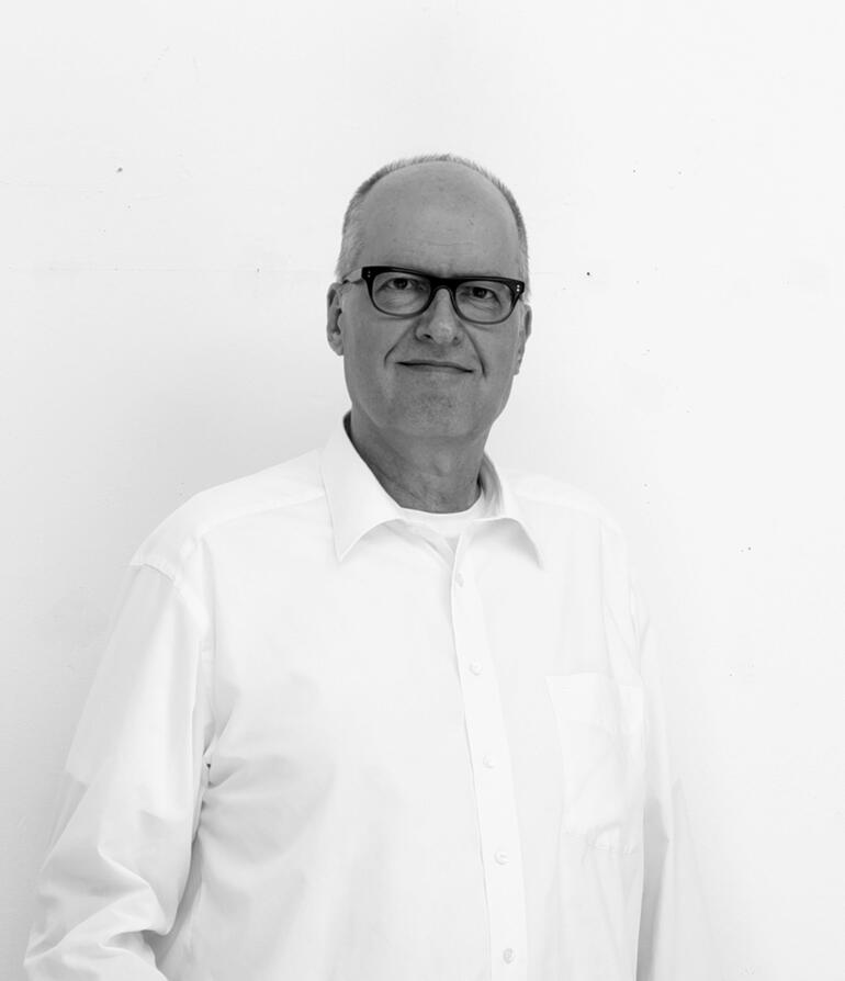 Jan Kolata Mann mit Brille lächelt vor einer weißen Wand, trägt ein weißes Hemd.