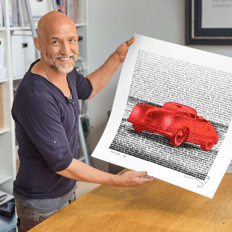 SAXA Mann hält einen Kunstdruck mit einem roten Auto vor einem Text hinter sich.